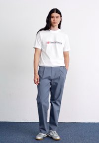 Witte katoenen t-shirt met een rood en zwart "New Balance" logo, gecombineerd met losse grijze broek en grijze sportschoenen, staand tegen een effen achtergrond.