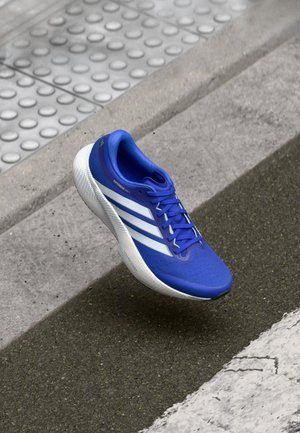 Blauer Laufschuh mit weißen Streifen und dicker Sohle, der über einen strukturierten Bordstein und eine Asphaltstraße schwebt.