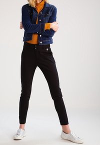 Person som står med armarna korsade, iklädd en blå denimjacka, orange polotröja, svarta byxor och vita sneakers mot en enhetlig bakgrund.