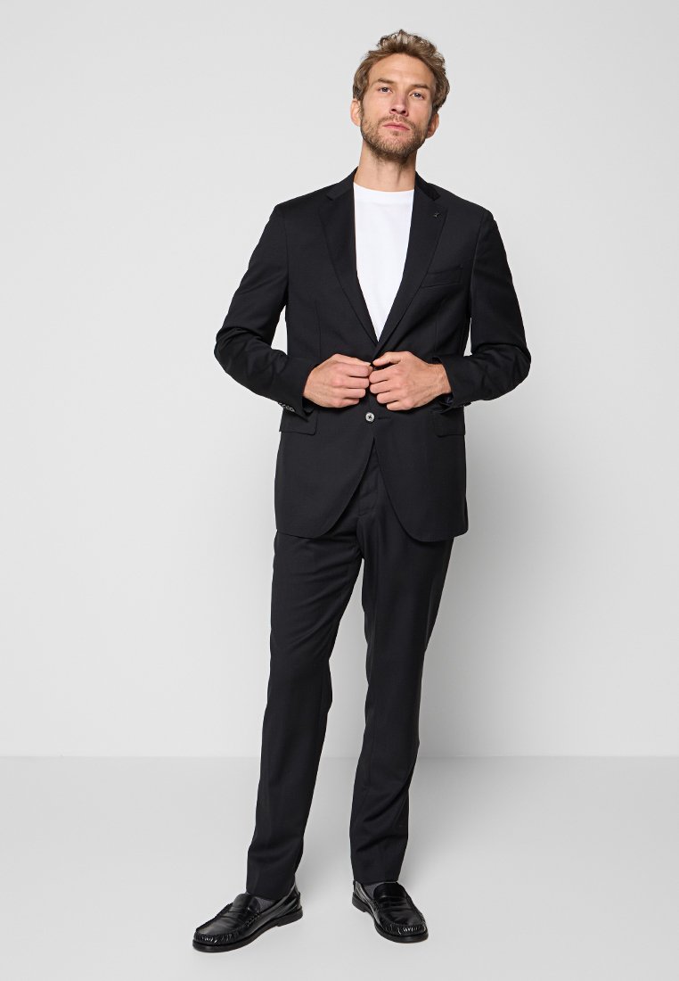 Man past een zwart colbert over een witte blouse aan, draagt bijpassende zwarte broek en zwarte instappers, staand tegen een effen grijze achtergrond.