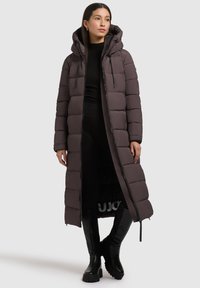 Dolga puhasta bunda temno rjave barve z zadrgo, nastavljivo kapuco in prešitim vzorcem. Kombinirana s črnimi škornji do kolen.