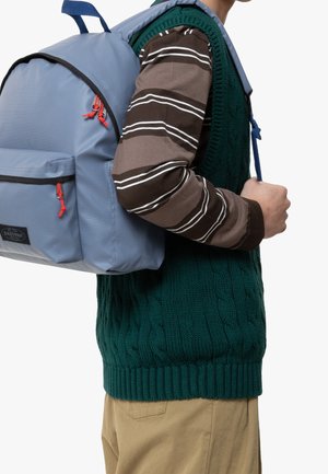 Osoba nosząca zieloną dzianą kamizelkę i brązową koszulę w paski, niosąca jasnoniebieski plecak z czerwonymi suwakami na jednym ramieniu.
