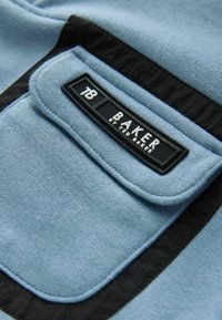 Niebieska tkanina o miękkiej fakturze, z czarnym akcentem i prostokątną metką oznaczoną "TB BAKER" z wytłoczonym napisem.