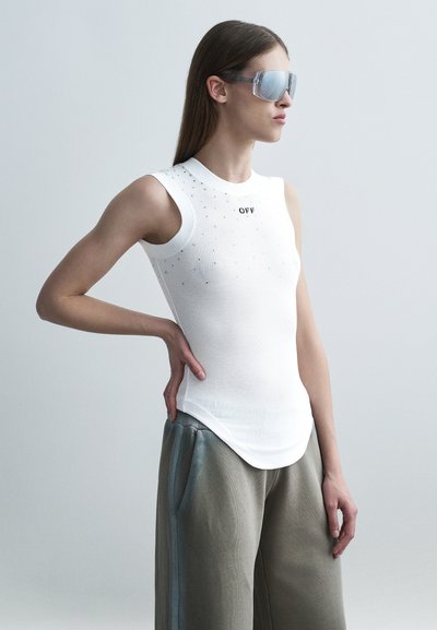 Femme portant un haut blanc sans manches avec le texte "OFF", un pantalon gris ample et de grandes lunettes de soleil réfléchissantes, debout avec la main sur la hanche.
