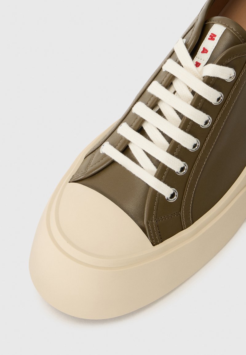 Gros plan sur une sneaker en cuir vert olive avec une épaisse semelle en caoutchouc beige, des lacets blancs et un texte rouge sur l'étiquette de la languette.