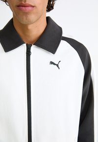 Puma jakna sa bijelim tijelom i crnim detaljima, rebrastim ovratnikom, zatvaranjem na patentni zatvarač i malim crnim logotipom na lijevoj strani prsa.