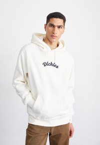 Sudadera blanca con capucha, que presenta un bolsillo frontal y el texto "Dickies" bordado en negro. El tejido parece suave con un ajuste relajado.