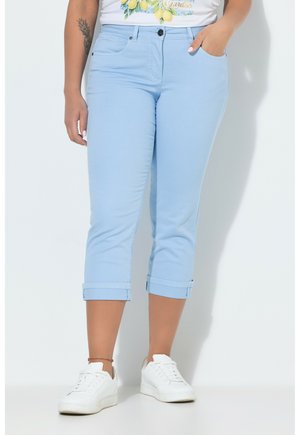 Donna che indossa pantaloni corti azzurri con orli risvoltati, sneakers bianche e maglietta bianca con stampa di limone, in piedi con una mano in tasca.