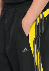 Melnas sporta bikses ar dzelteniem un pelēkiem akcentiem, ar elastīgu jostu, sānu kabatām un Adidas logo tuvumā apakšmalai.