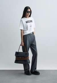 Bílé tričko s nápisem "MOSCHINO Milano" v černé barvě, šedé kalhoty s vysokým pasem, nadměrně velká šedá taška s hnědým popruhem a robustní černé boty.