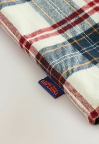 Ruudullinen kangas, jossa on punaisia, sinisiä ja kermaisia värejä. Pehmeä tekstuuri, jossa näkyy oranssi "Superdry" -merkki sinisellä taustalla.