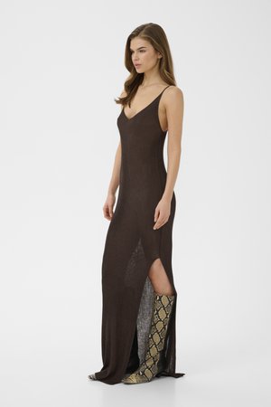 Kvinde iført en ærmeløs mørkebrun maxikjole med sideslids, kombineret med knæhøje støvler med slangeprint, stående sideværts på hvid baggrund.