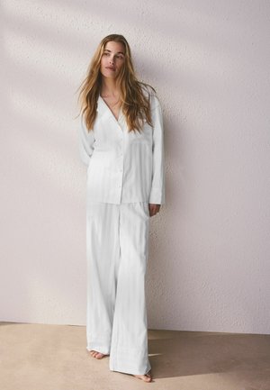 Woman with long hair wearing loose white button-up shirt and wide-leg pants, standing barefoot against a textured pale pink wall.