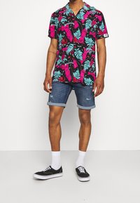 Korte mouwen shirt met een zwarte basis en levendige roze, blauwe en groene tropische patronen. Draagt bijpassende denim shorts en zwarte sneakers.