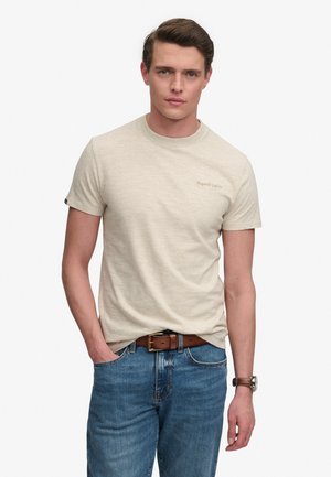 Man draagt een beige Superdry t-shirt met korte mouwen, blauwe spijkerbroek vastgemaakt met een bruine riem en een bruin polshorloge, staand met één hand in de broekzak.
