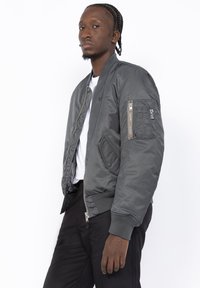 Giacca bomber grigia con una texture liscia, chiusura frontale con zip, polsini a coste e una tasca con zip sulla manica. Indossata sopra una T-shirt bianca.