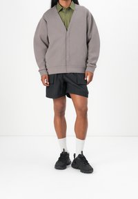 Šedý cardigan s výstřihem do V, černé šortky a černé tenisky. Oblečení má oversized střih. Bílé ponožky viditelné nad teniskami.