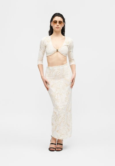 Γυναίκα με σκούρα βρεγμένα μαλλιά, φοράει κεχριμπαρένια γυαλιά ηλίου, ένα μπεζ crop top με μοτίβα και μανίκια μέχρι τον αγκώνα, μια μακριά φούστα με ψηλή μέση που ταιριάζει και μαύρες ψηλοτάκουνες γόβες.