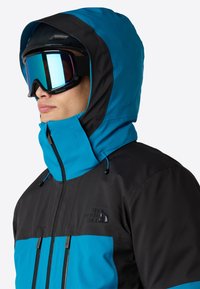 Blå och svart skidjacka med hög krage, fickor med dragkedja och justerbar huva; har en logotyp på bröstet och en slät textur.