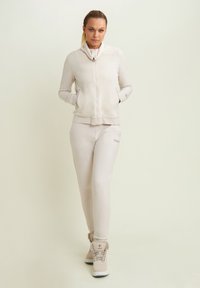 Giacca zip beige chiaro con collo a imbuto e due tasche, abbinata a pantaloni tuta coordinati con dettagli del logo. Sneakers stile stivaletto completano il look.