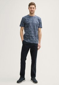 Navy blue short-sleeve shirt with a textured pattern, paired with dark pants and athletic shoes with a white sole, standing against a neutral background.