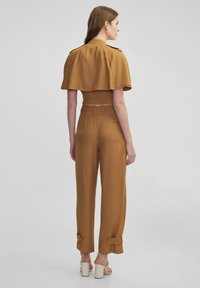 Cropped jacket and high-waisted trousers in mustard fabric. Jacket features a cape design with buttons, trousers have rolled cuffs and button details.