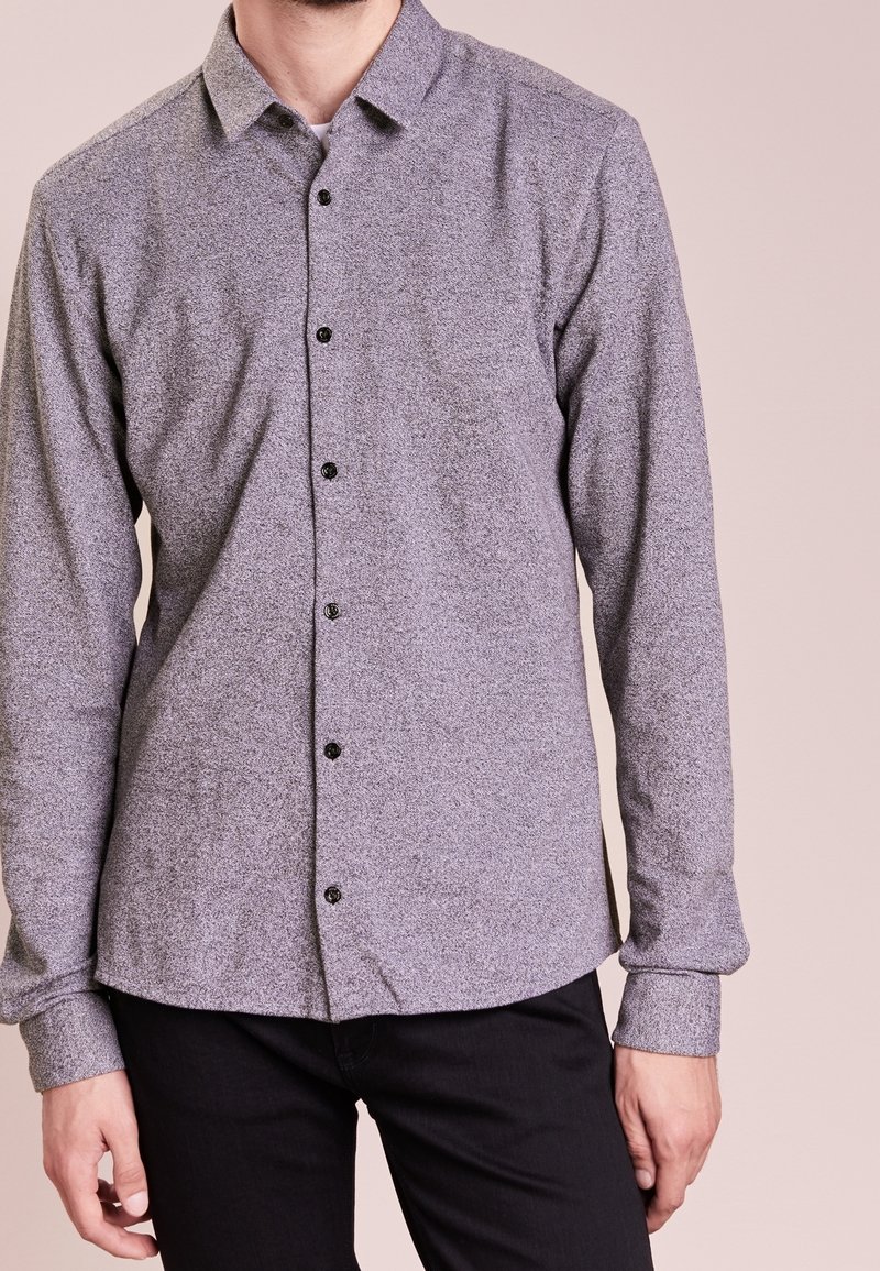 Mand iført en langærmet, knaplukket grå skjorte med et subtilt mønster og sorte bukser foran en ensartet lys baggrund.