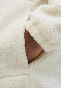 Tessuto in pelliccia sintetica color crema con una superficie texturizzata e pelosa. Un braccio marrone è visibile attraverso un'apertura nella manica dell'indumento.