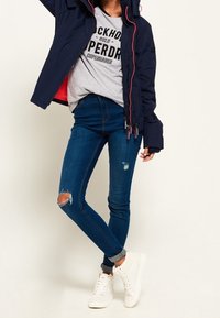 Marine blå puffjakke med pink foring, grå grafisk t-shirt, slidte blå jeans og hvide sneakers.