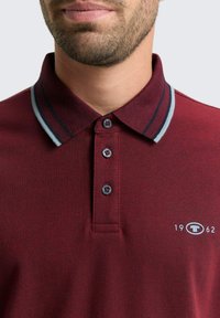 Burgunderfarbener Poloshirt aus strukturiertem Stoff, mit einem Kragen mit zwei Knöpfen und kontrastierenden grauen und marineblauen Streifen am Kragen. Mit einem Logo auf der Brust.