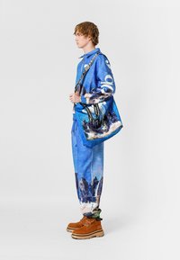 Conjunto azul texturizado que incluye una chaqueta con cremallera y pantalones con estampados de montañas. Botas marrones y un bolso de hombro con patrón completan el look.