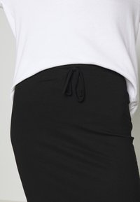 Gros plan sur une personne portant un pantalon noir avec un cordon de serrage noué à la taille et un haut blanc rentré.