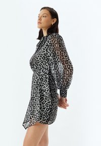 Korte zwart-wit luipaardprint jurk met lange, transparante mouwen en een getailleerde taille. Rond aflopende zoom en knoopsluitingen bij de manchetten. Subtiele glans in de stof.
