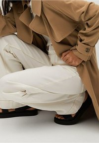 Beiger Trenchcoat mit breiten Revers, kombiniert mit cremefarbenen, weit geschnittenen Hosen und schwarzen Plattform-Sandalen mit strukturierten Riemen.