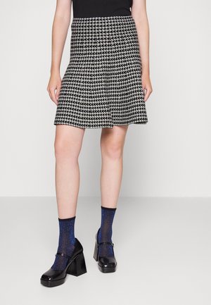 Femme portant une jupe en pied-de-poule noir et blanc, un haut noir, des chaussettes étincelantes bleues et des chaussures à plateforme Mary Jane noires, se tenant devant un fond blanc.