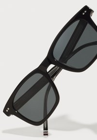 Gafas de sol negras con montura cuadrada, lentes oscuros y detalles plateados. Las varillas presentan un patrón a rayas en rojo, blanco y azul.