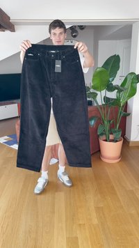 Juoda veliūro kelnės su plačiomis kojomis, turinčios mygtuką ir užtrauktuką, priekinėmis kišenėmis ir prisegamu etiketė. Tekstūruotas audinys.