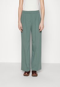 Pantalons à jambes larges en tissu vert doux, avec des plis avant, une taille lisse, associés à des sabots marron.