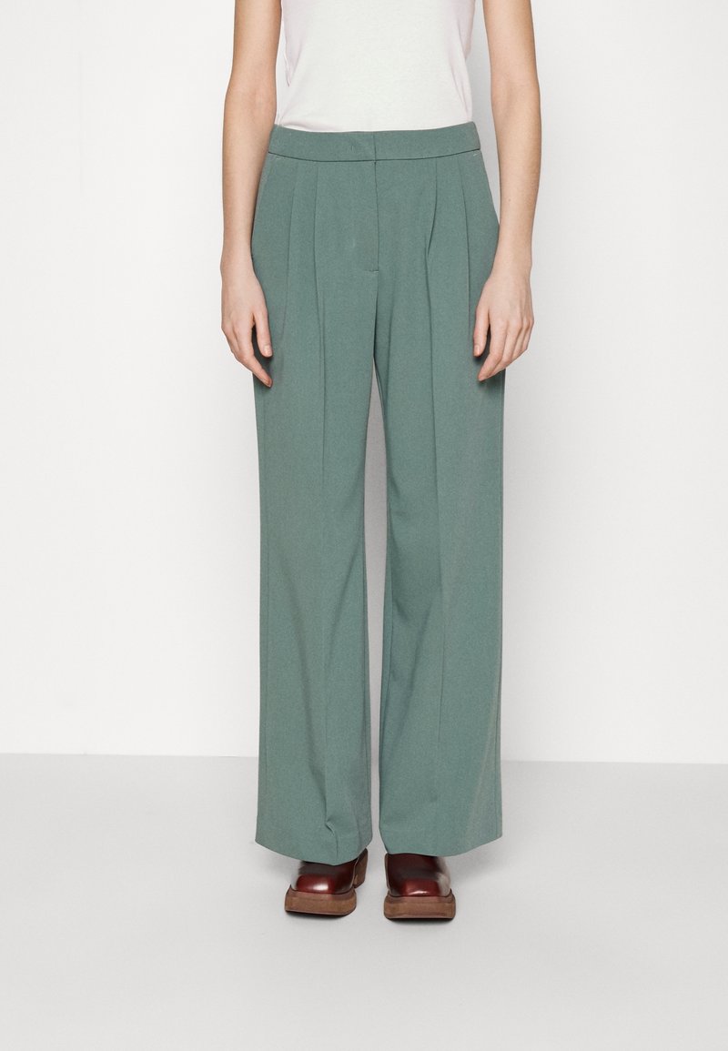 Pantalons à jambes larges en tissu vert doux, avec des plis avant, une taille lisse, associés à des sabots marron.
