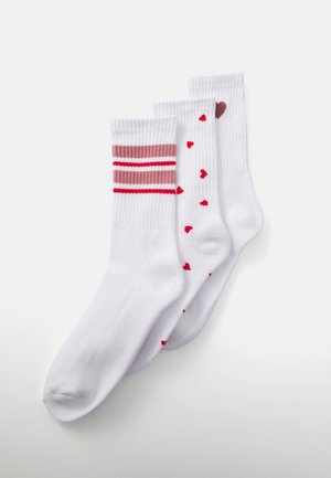 Three white crew socks, one with pink and red stripes, one with red hearts, and one with a single brown heart near the cuff.