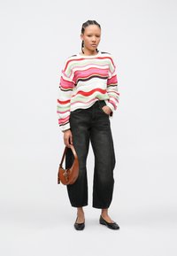 Γυναίκα με πλεξούδες, φοράει πολύχρωμο ριγέ πουλόβερ, μαύρο τζιν, μαύρες μπαλαρίνες, κρατώντας μια καφέ τσάντα, μπροστά σε ένα απλό λευκό φόντο.