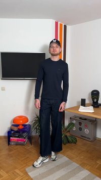 Langärmliges dunkelblaues Hemd, dunkle, weit geschnittene Jeans und graue Sneakers. Steht auf einem hellen Teppich neben einer bunten Lampe und einem Holztisch.