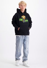 Zwarte hoodie met een cartoonafbeelding van een aap in een groene autothema-outfit, gecombineerd met lichtblauwe versleten spijkerbroeken en witte sneakers.