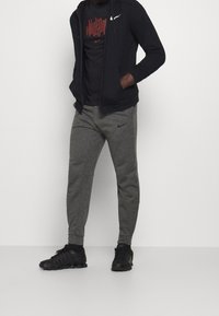 Sudadera negra con cremallera y logo blanco, combinada con pantalones de chándal gris oscuro que presentan un pequeño logo, y zapatillas deportivas negras. Textura de tela suave.