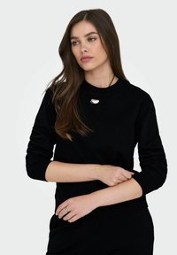 Γυναίκα με μακριά καστανά μαλλιά, φορώντας μαύρο μπλουζάκι με μακριά μανίκια και ασημένιο κολιέ, που ρυθμίζει το αριστερό της μανίκι με ουδέτερη έκφραση.