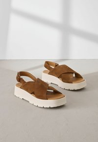 Sandali platform in suede marrone con design a strappo incrociato, suole in gomma beige e cinturini alla caviglia regolabili. Parte superiore testurizzata con un plantare liscio.