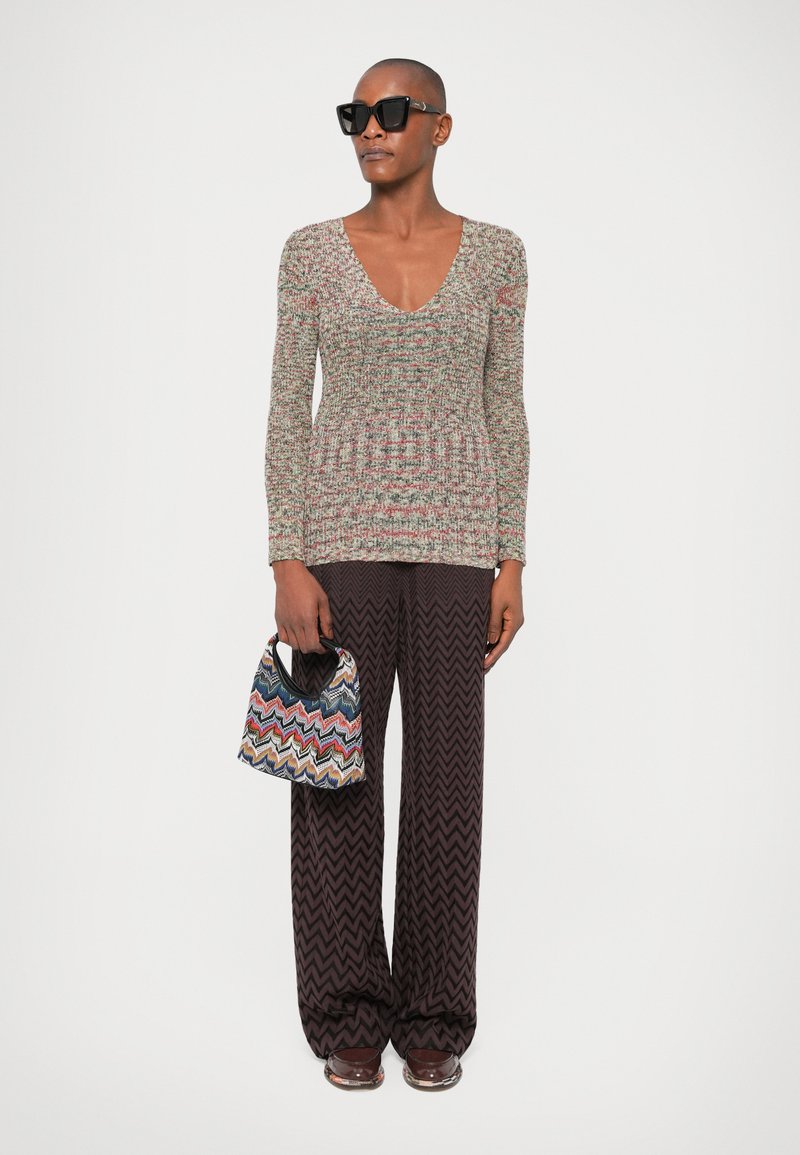 Bunt gestricktes Oberteil mit tiefem V-Ausschnitt, kombiniert mit dunkelbraunen, weit geschnittenen Hosen im Zickzackmuster. Einschließlich einer kleinen, bunten Handtasche.