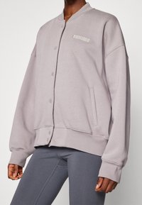 Giacca bomber grigio chiaro in tessuto morbido, con chiusura a scatto, tasche laterali e un logo ricamato discreto sul petto.