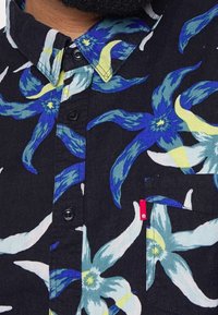 Chemise noire à manches courtes avec un motif floral en bleu, blanc et jaune, col boutonné et une poche poitrine avec une étiquette rouge.