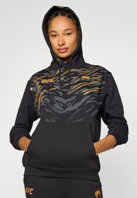 Sweatshirt preta com bolso canguru, apresentando um padrão geométrico em cinza e laranja, capuz com cordão e logótipo "UFC" na frente.